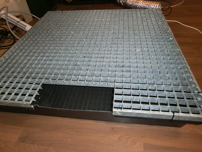 Ein Gitterrost über einer schwarzen Auffangwanne, der auf einem Holzboden liegt. Daneben sind ein Lüftungsschlauch und Kabel zu sehen.