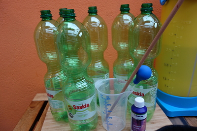Mehrere leere Plastikflaschen stehen auf einer Holzoberfläche vor einer orangefarbenen Wand. Daneben befinden sich ein Messbecher mit einem Holzstab und einer Pipette sowie eine kleine Flasche mit der Aufschrift "Plagron Power Roots".
