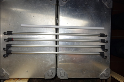 Vier Aluminiumstangen mit schwarzen Haken an den Enden, daneben eine weiße Kunststoffstange, liegen auf einer metallischen Oberfläche.
