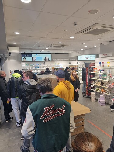 Eine Gruppe von Menschen steht in einer Schlange in einem Geschäft, das Kosmetikartikel verkauft. Im Vordergrund trägt eine Person eine Jacke mit der Aufschrift "Kani est 1989".
