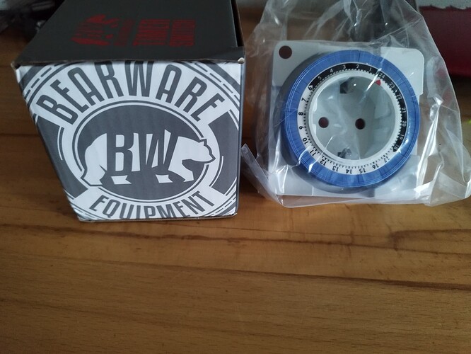 Eine Schachtel mit der Aufschrift "BEARWARE EQUIPMENT" und einem Bärenlogo daneben, sowie eine blaue mechanische Zeitschaltuhr in einer Plastikverpackung.