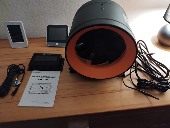 Ein Abluftventilator mit orangefarbenem Rand, ein Smart Controller und ein Thermometer/Hygrometer liegen auf einem Holztisch.