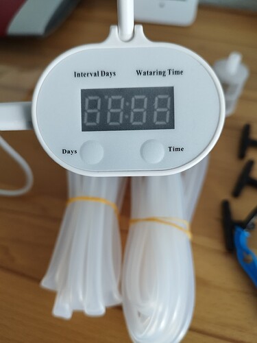 Nahaufnahme eines automatischen Bewässerungssystems mit einem digitalen Display, das "88:88" anzeigt, und mehreren Schläuchen, die mit gelben Gummibändern gebündelt sind.