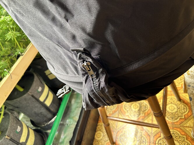 Nahaufnahme eines schwarzen Kleidungsstücks mit Reißverschluss, im Hintergrund sind Pflanzen und Growbags zu sehen.