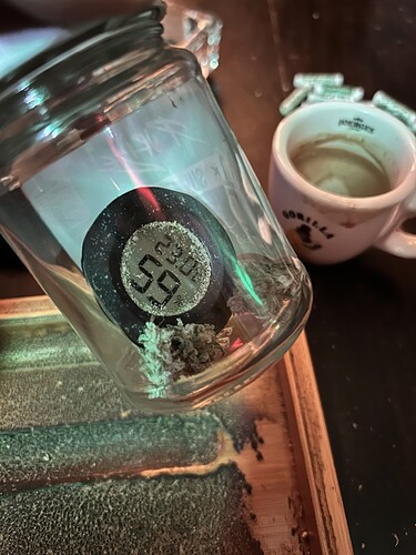 Ein Glas mit Cannabis-Buds und einem Hygrometer, das eine Luftfeuchtigkeit von 59% anzeigt, neben einer Kaffeetasse.