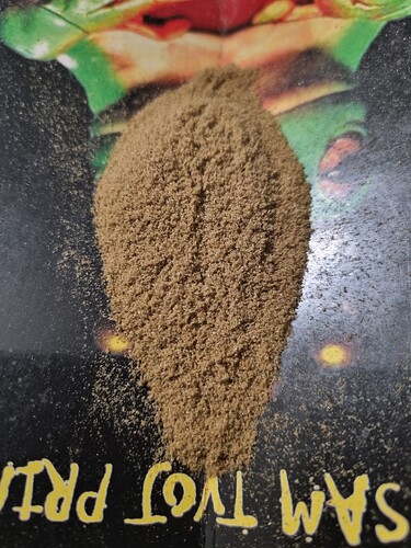 Ein Haufen feines, hellbraunes Pulver liegt auf einer schwarzen Oberfläche. Im Hintergrund sind bunte, schemenhafte Bilder zu erkennen. Am unteren Bildrand sind gelbe Buchstaben zu sehen, die auf dem Kopf stehen und "SAM TVOT PR" lauten.