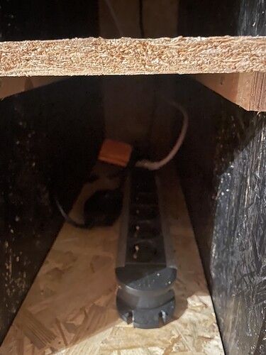 Eine Mehrfachsteckdose mit mehreren Anschlüssen und einem orangefarbenen Stecker, die in einem dunklen Schrank auf einer Holzplatte liegt.