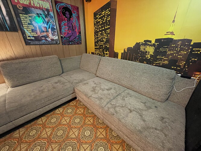 Ein Ecksofa steht auf einem gemusterten Teppichboden vor einer Wand mit Holzvertäfelung. An der Wand hängen zwei Poster: eines ist ein Filmplakat für "Pulp Fiction" mit einem leuchtend grünen Cannabis-Symbol, das andere zeigt eine stilisierte Frau. Daneben ist eine Wandtapete mit einer Skyline bei Nacht im gelben Licht angebracht.