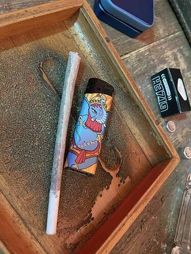 Ein Joint liegt neben einem Feuerzeug mit Elefantenmotiv auf einem Holzteller. Daneben befindet sich eine kleine Schachtel mit Zigarettenpapier und ein paar Filtern.