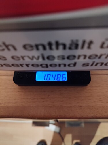 Eine digitale Waage zeigt 104.86g an, darüber ein unscharfer Text auf einem roten Hintergrund.