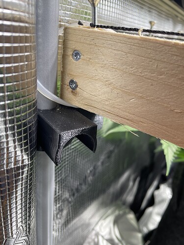 Nahaufnahme einer 3D-gedruckten Halterung, die an einem vertikalen Rohr befestigt ist und ein Holzbrett stützt, mit Cannabisblättern im Hintergrund.