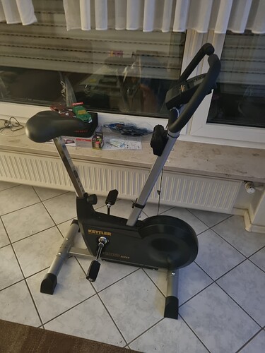 Ein schwarzes Kettler Heimtrainer-Fahrrad steht auf einem gefliesten Boden vor einem Fenster.
