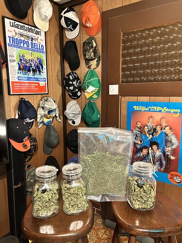 Drei Gläser und ein Plastikbeutel gefüllt mit getrockneten Cannabis-Buds, daneben ein Poster der Band "Hans and the Hit Singers" und eine Sammlung von Baseballkappen an der Wand.