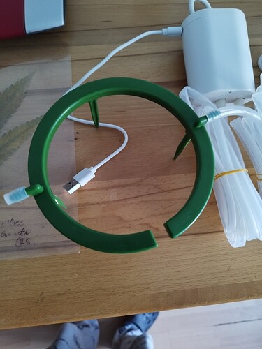 Ein grüner Bewässerungsring mit angeschlossenen Schläuchen und einer weißen Pumpe im Hintergrund, die über ein USB-Kabel aufgeladen wird.