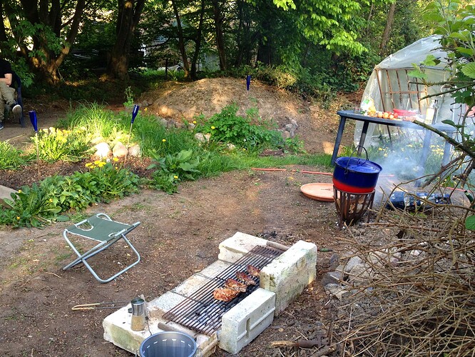 Ein Gartenbereich mit einem provisorischen Grill aus Betonsteinen, auf dem Fleisch gegrillt wird, sowie einem Topf auf einer Feuerstelle und einem kleinen Klappstuhl.